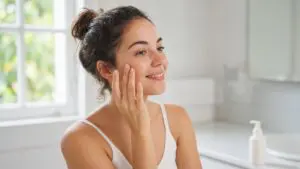 Smiling woman in her late 20s pressing serum into her cheek in a bright morning bathroom with a pump bottle on the counter, part of her vitamin c serum for 20s routine