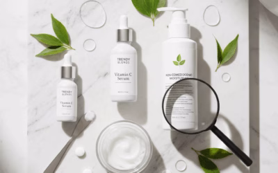 A top-down view of various skincare products on a white marble surface. Key items include two white bottles of "TRENDY BLENDS Vitamin C Serum" with droppers, a larger white pump bottle prominently labeled "NON-COMEDOGENIC MOISTURIZER," and a white jar of face cream with a small spatula beside it. A magnifying glass is placed over the non-comedogenic moisturizer bottle, suggesting scrutiny and informed choice. Several green leaves and clear water droplets or serum beads are scattered around, adding a fresh and natural feel.