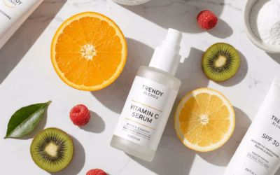 A flat lay of Trendy Blends skincare products, featuring a white bottle of Vitamin C serum as the centerpiece. Surrounding it are fresh fruit slices of orange, lemon, and kiwi, along with raspberries, emphasizing the natural, antioxidant-rich ingredients. Other Trendy Blends products like a moisturizer and SPF are also visible on a clean white marble surface.
